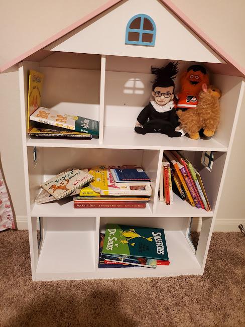 A children's bookcase in the shape of a dollhouse. Visible are several books be Dr. Seuss, including "Hop on Pop" and "The Sneetches", a few by Leslie Patricelli, such as "Mad, Mad, Mad", and "A is for Awesome", by Eva Chen, an alphabet book featuring inspiring women. Also visible on the shelves are three dolls: one of an alpaca, one of Philadelphia Flyers mascot Gritty, and one of Ruth Bader Ginsberg.