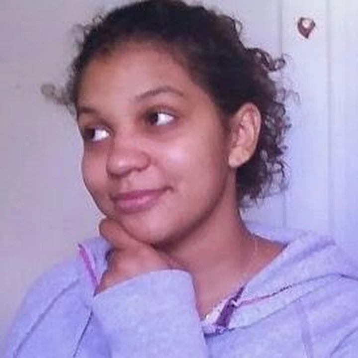 Photograph from the chest up. Biracial woman in her mid twenties. She has curly hair, which is pulled back. She is wearing a purple hoodie.
