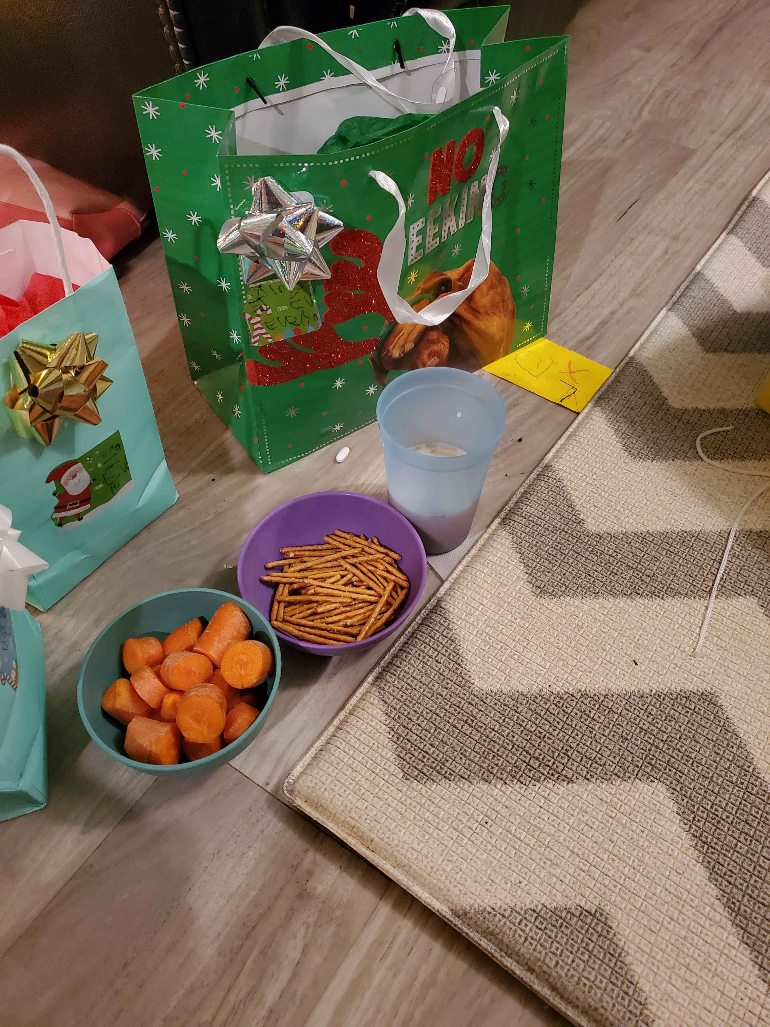 A couple Christmas presents in bags on the floor. In front of them are two bowls and a cup. The bowl on the left is sea blue and contains several carrot slices. The bowl in the middle, colored purple, curtains pretzel sticks. The cup on the right is blue and half full. The contained liquid isn't visible.