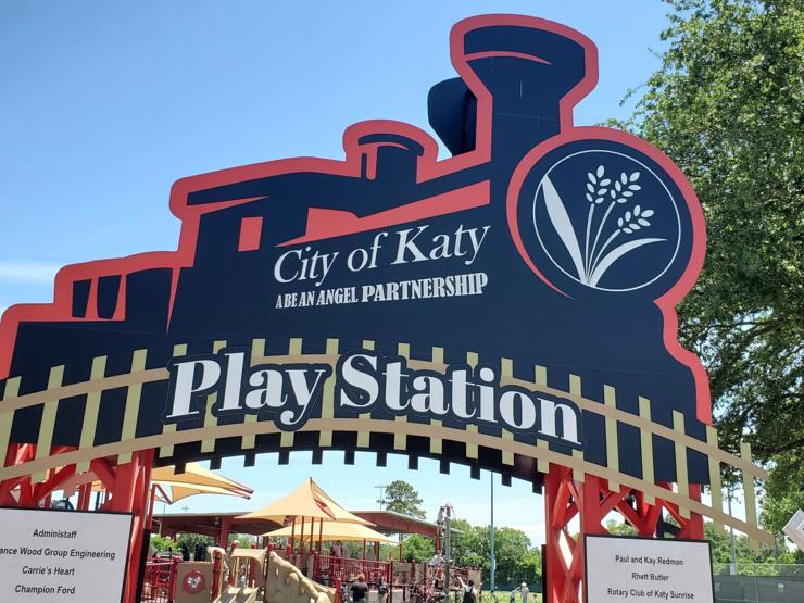 Sign in front of the entrance of a playground. The sign is shaped like a train and train tracks. On the side of the train are the words "City of Katy" and underneath "A Be an Angel Partnership", although due to poor kerning, it looks like "Abe an Angel". Written along the train tracks beneath the train are the words "Play Station."