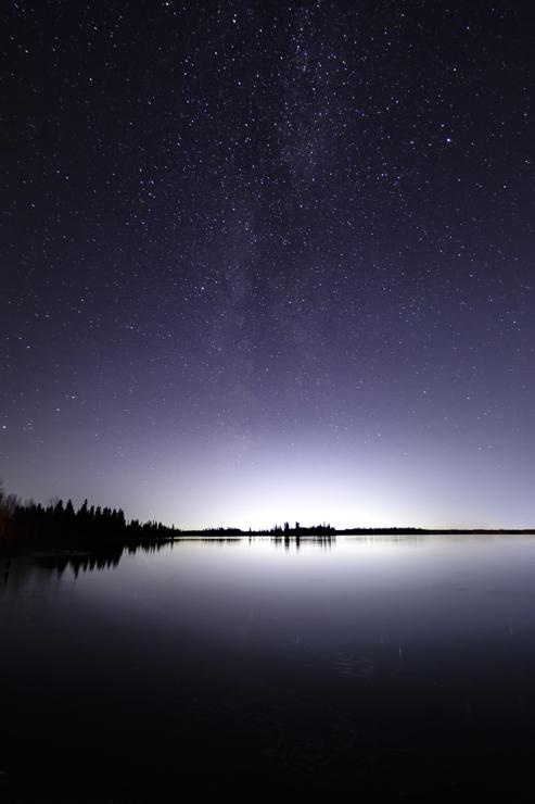 A night sky fades from black to purple to white along the horizon, filled with countless silver stars. Along the straight line demarcating earth from sky, a forest slopes down slightly from left to centre, standing out barely against the distant glow. Below, a still lake reflects back the forest and horizon’s light. 