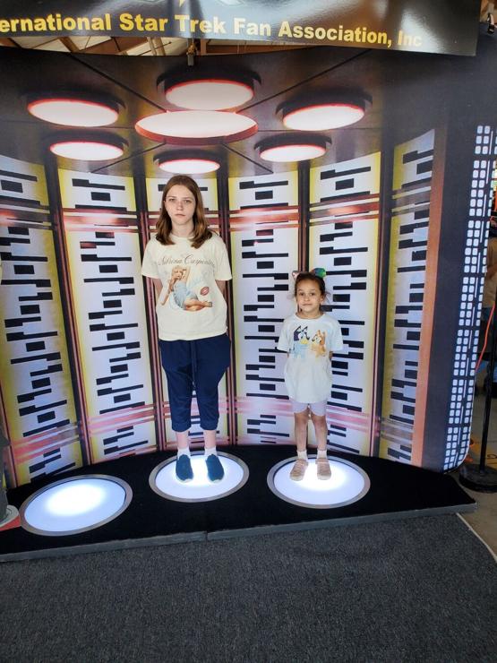 Two girls, one teenage and one preschooler, standing on the transporter from Star Trek: The Original Series.
Both have a very stoic, Vulcan look on he their faces.
