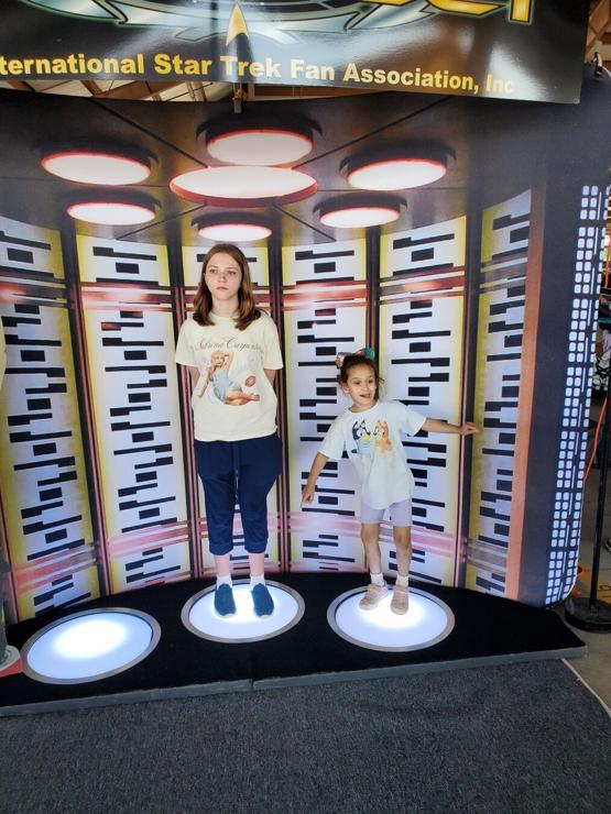 Two girls, one teenage and one preschooler, standing on the transporter from Star Trek: The Original Series.
The preschooler, on the right, is holding her hands out and smiling. The teenager, in the middle, has a very stoic, Vulcan look on her face