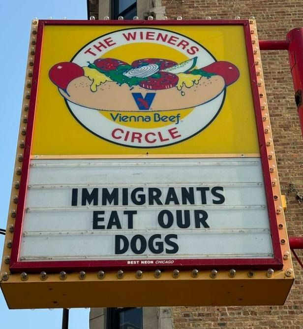 A photo of The Wieners Circle sign in Chicago.  The top is a Vienna Beef hot dog with a yellow background.  The bottom says:  "IMMIGRANTS EAT OUR DOGS"
