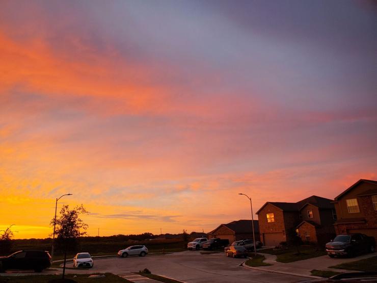 A cul-de-sac with a couple houses and cars visible. There are cirrus clouds visible, shining orange and red against a purple-ish blue sky. The houses are orange and pink, reflecting the light of the setting sun.