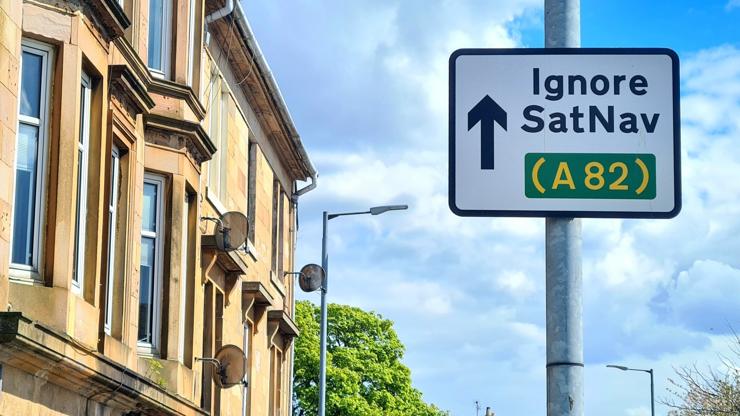 A offical road sign in Bowling near Glasgow with an arrow pointing straight ahead accompanied by the words Ignore SatNav (A82).