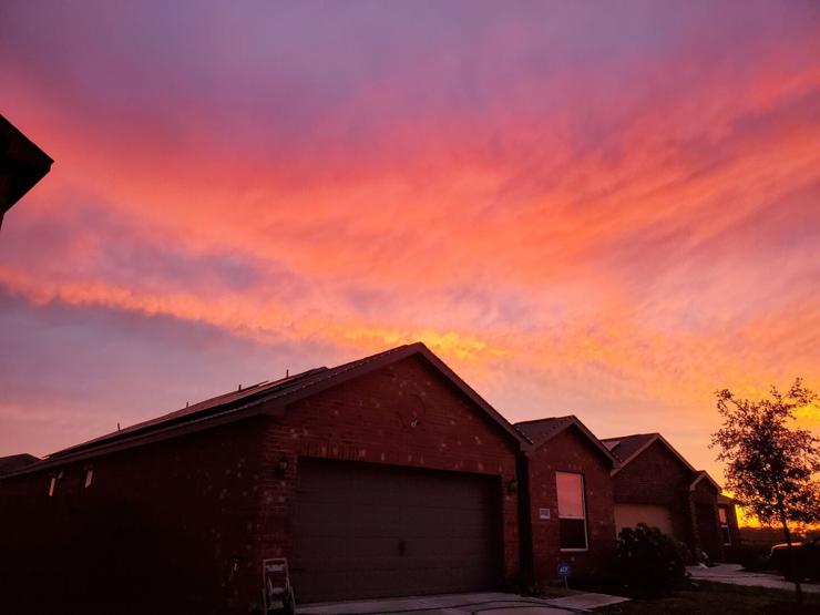 A cul-de-sac with a couple houses and cars visible. There are cirrus clouds visible, shining pink against a purple-ish blue sky. The houses are dark purple, reflecting the light of the setting sun. There is a small tree on the right side with bright, almost neon, orange clouds visible behind it.