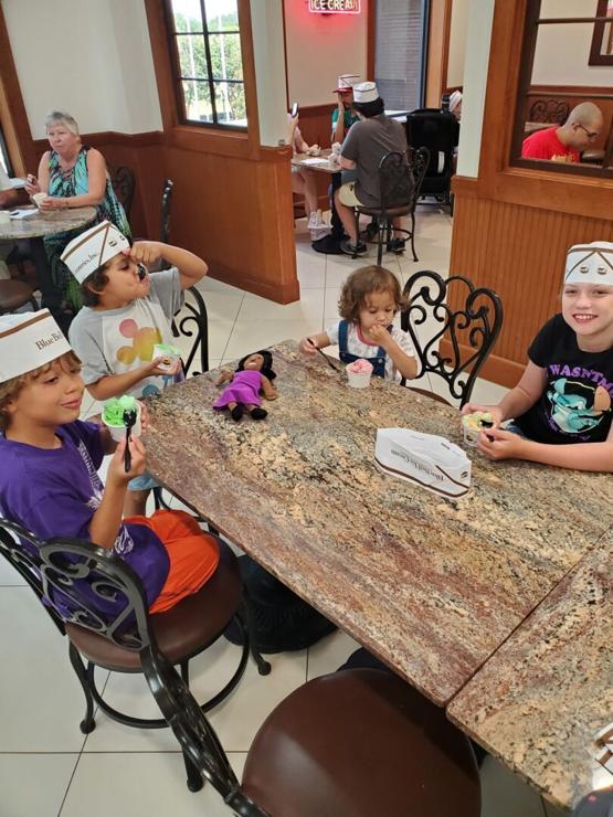 Four children of various ages sit around a table eating ice cream. A doll with the likeness of Michelle Obama sits on the table on front of a toddler girl.