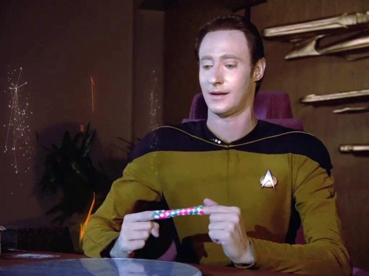 Data is sitting at a table in a high back conference type chair. Some model starships are decorating the wall behind him. He is looking down with a dumb half smile and has one finger each in a finger trap toy that is colored pink and green. 