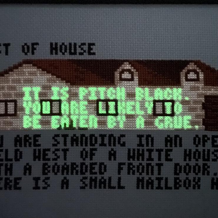 Zoomed-in “West of House” cross stitch in dim light. In green glowing letters: “IT IS PITCH BLACK. YOU ARE LIKELY TO BE EATEN BY A GRUE.”