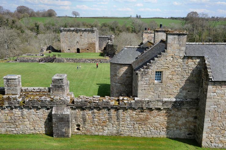 Craignethan Castle in Lanarkshire. The image shows a view down on to a castle that comprises a stone wall of modest height cutting across the foreground with a harge old stone house at its right-hand end. Beyond the wall and house is a mown grassy area, then a ditch crossed by a footbridge and then a squat tower house also made from honey coloured stone. Beyond is are threes then distant farmland. The castle is in sunlight.