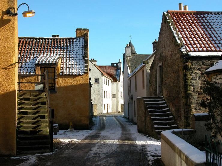 Culross in Fife. The image shows a lane leading between very old buildings towards a white three-storey building in the distance. The buildings on the left are coated in a yellow harling, while those on the right are finished in natural stone and white. The roofs are mainly red pantiles. There are open stone stairways leading up to the first floor on both sides of the lane. There are signs of snow on the ground and the sky is blue.