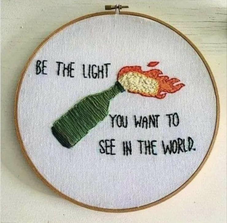 A cross stitch project showing a green glass bottle diagonal as if in motion. A flame erupts from the mouth. Black block text reads, "be the light you want to see in the world."