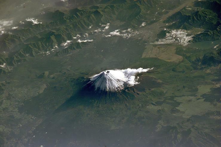 A snow-capped stratovolcano surrounded by green fields.