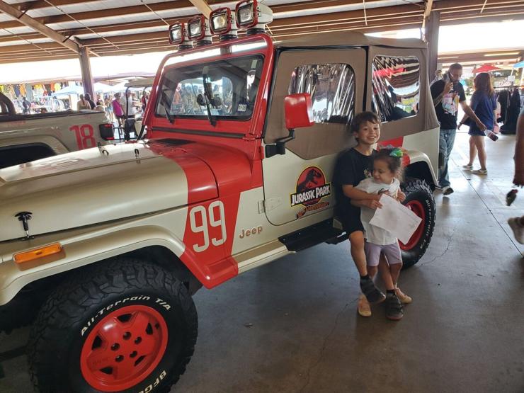 A 9-year-old boy wearing a black T-shirt and a preschool aged girl wearing a rainbow boy in her hair stand in front of the Jeep from Jurassic Park. The boy is hugging the girl from behind, in a loving, brotherly way.