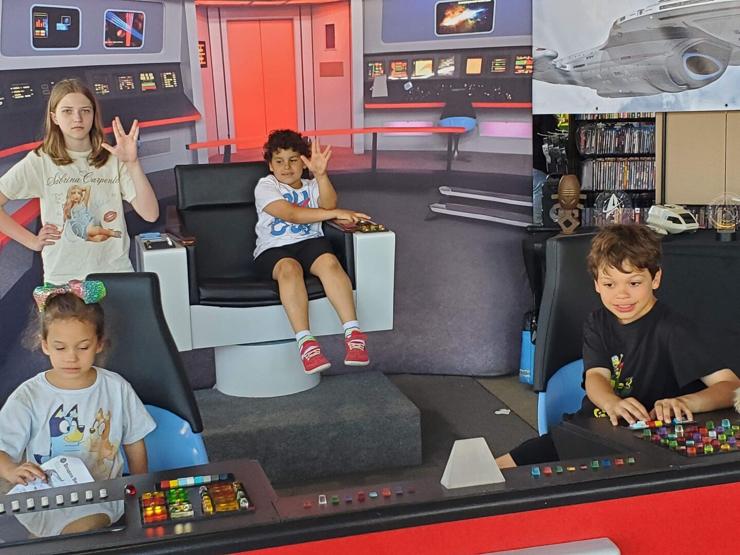 Four children on the bridge of the USS Enterprise from Star Trek. In the captain's is a little boy playing with the controls and holding up a Vulcan salute high. Standing next to him, with a Vulcan expression in her face, is a teenage girl, wearing a Sabrina Carpenter T-shirt, also doing a Vulcan salute. In Sulu's chair is a preschool aged girl wearing a Bluey T-shirt and a rainbow bow. In Chekov's chair is a boy, older than the captain, working the controls.