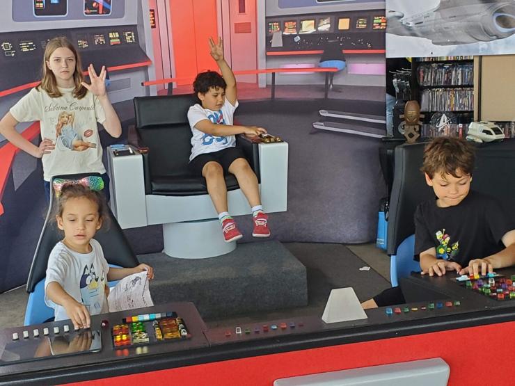 Four children on the bridge of the USS Enterprise from Star Trek. In the captain's is a little boy playing with the controls and holding up a Vulcan salute high. Standing next to him, with a Vulcan expression in her face, is a teenage girl, wearing a Sabrina Carpenter T-shirt, also doing a Vulcan salute. In Sulu's chair is a preschool aged girl wearing a Bluey T-shirt and a rainbow bow. In Chekov's chair is a boy, older than the captain, working the controls.