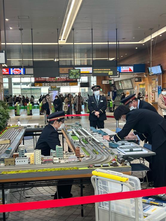 Inside the ticket gates (which can be seen in the background) four men in train station uniforms are setting up a large model train set. 
