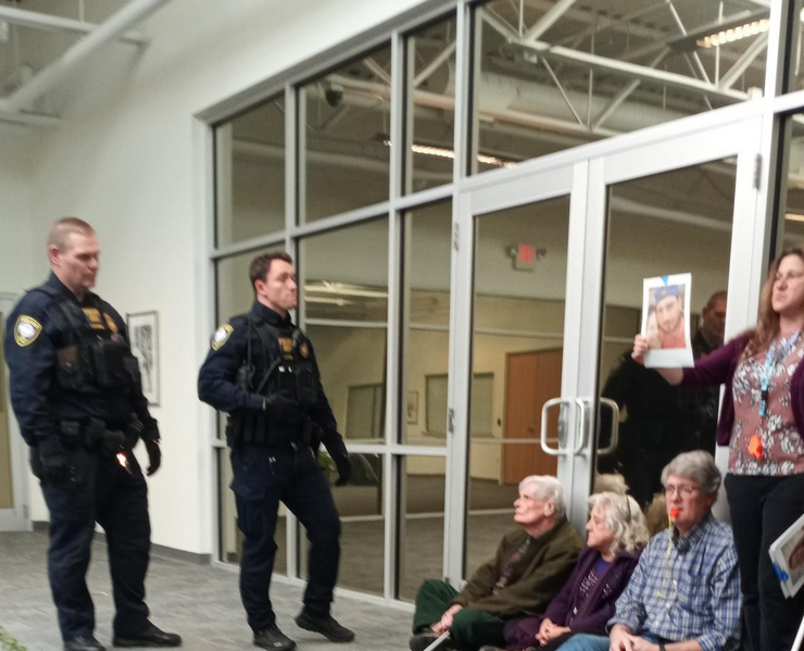 Three white/gray haired people sit on the floor in front of a glass door, some with whistles in their mouths. Next to them stands a younger woman holding a photo, presumably of a person killed in ICE custody. Off to the left two cops watch them.