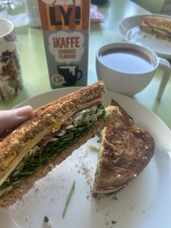 Two-tier toasted sandwich cut in half. It’s filled with hummus, greens, vegan cold cuts, mustard and cheese. Next to it there’s a large cup of coffee with oat milk. 