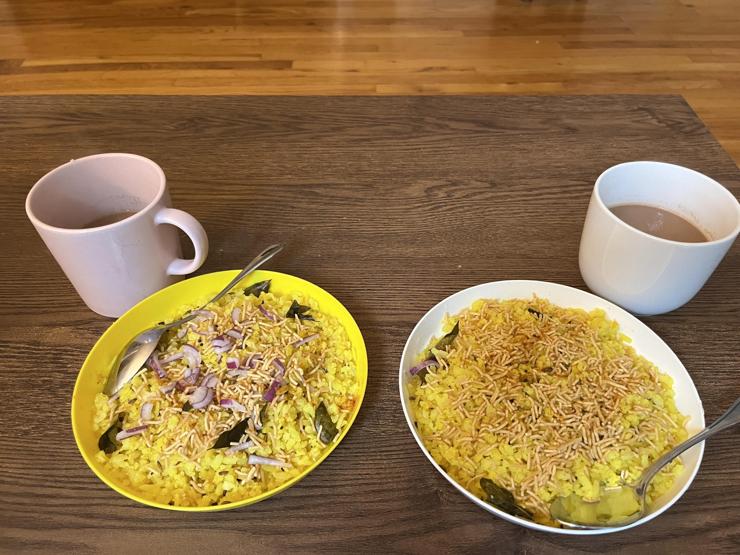 The image features two bowls of poha garnished with onions and crispy noodles, placed on a wooden table. There are two cups of tea beside the bowls, one pink and one white.
