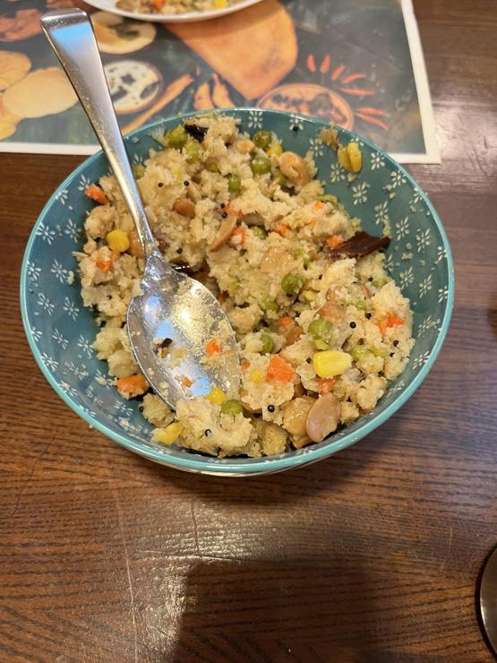A bowl contains a mixture of coarse grains, vegetables, and nuts, featuring peas, corn, carrots, and spices. A spoon is resting in the bowl, which is placed on a wooden table with a colorful placemat in the background.