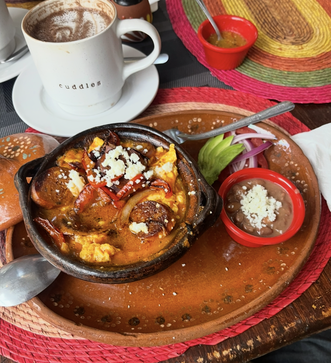 A cast iron dish with sausage and vegetable and egg goodness in it. On a plate with beans and garnish. A white mug of hot chocolate in the upper right. 