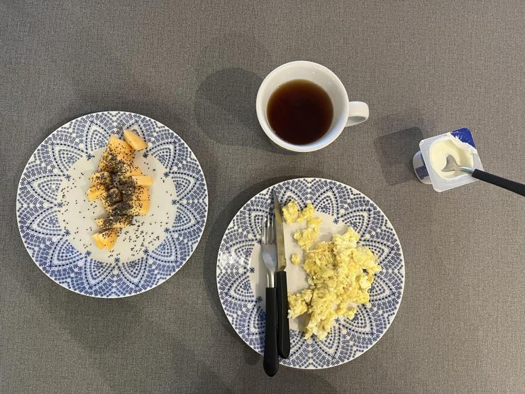 A breakfast spread featuring two patterned plates. One plate contains diced mango and chia seeds, while the other has scrambled eggs. A cup of tea sits nearby, alongside a small cup of yogurt with a spoon. The table is a neutral gray.