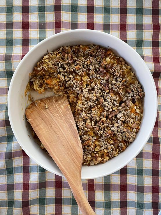 A plan view of the whole oven dish of oven porridge, after cooking. It’s a white Arabia oven dish, maybe 25 cm across. A portion has been removed and a wooden spatula is lying in the resulting gap. The tablecloth in the background is red, green and white checked pattern.
