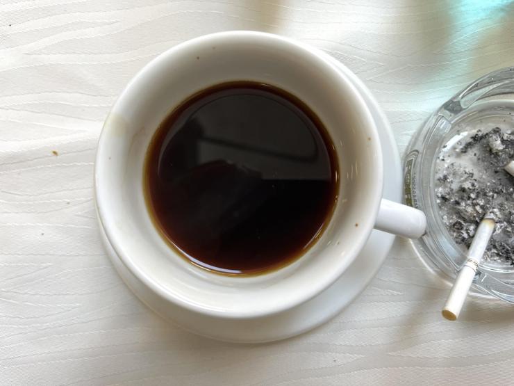 Photo looking down at a cup of black coffee, and next to it a half-smoked cigarette resting in an ashtray